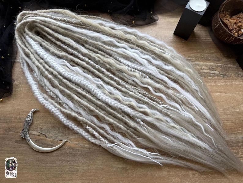 a collection of long, white braided hair strands arranged on a wooden surface. There is also a small metal object, possibly a hair clip, placed next to the hair strands.