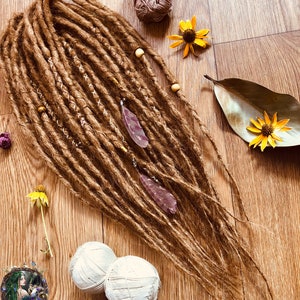 K&ouml;nnte beinhalten: Braune Dreadlocks mit Holzperlen und lila Federakzenten. Die Dreadlocks sind auf einer Holzoberfl&auml;che mit zwei Kn&auml;ueln wei&szlig;er Wolle, Trockenblumen und einem Logo mit der Aufschrift "Dreads Forest" angeordnet.