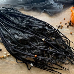 May include: Black dreadlocks with wavy ends, adorned with wooden and white beads. The dreads are arranged on a wooden surface, with a pumpkin-shaped container and decorative beads in the background. The logo "Dreads FOREST" is visible.