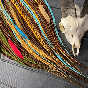 May include: A collection of colorful synthetic dreadlocks in shades of brown, yellow, green, blue, and red. The dreadlocks are arranged in a fan shape and are accented with feathers.