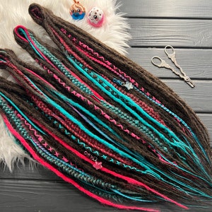 May include: Dark brown dreadlocks with vibrant pink and turquoise accents. The dreads are decorated with intricate stitching and small charms. The image also includes a pair of decorative scissors and small figurines.