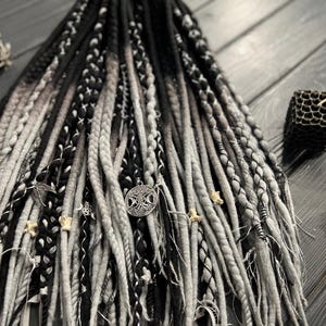 Könnte beinhalten: Ein Set aus schwarzen und grauen synthetischen Dreadlocks mit silbernen und knochenfarbenen Perlen. Die Dreadlocks sind fächerförmig angeordnet und haben verschiedene Texturen.