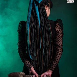 Handcrafted synthetic dreadlock extensions in forest green and black ombre. The set features a mix of thin twisted dreads, classic braids, and fishtail braids arranged in a fan shape. A model in a gothic setting holds a black skull, emphasizing a mystical forest witch aesthetic.