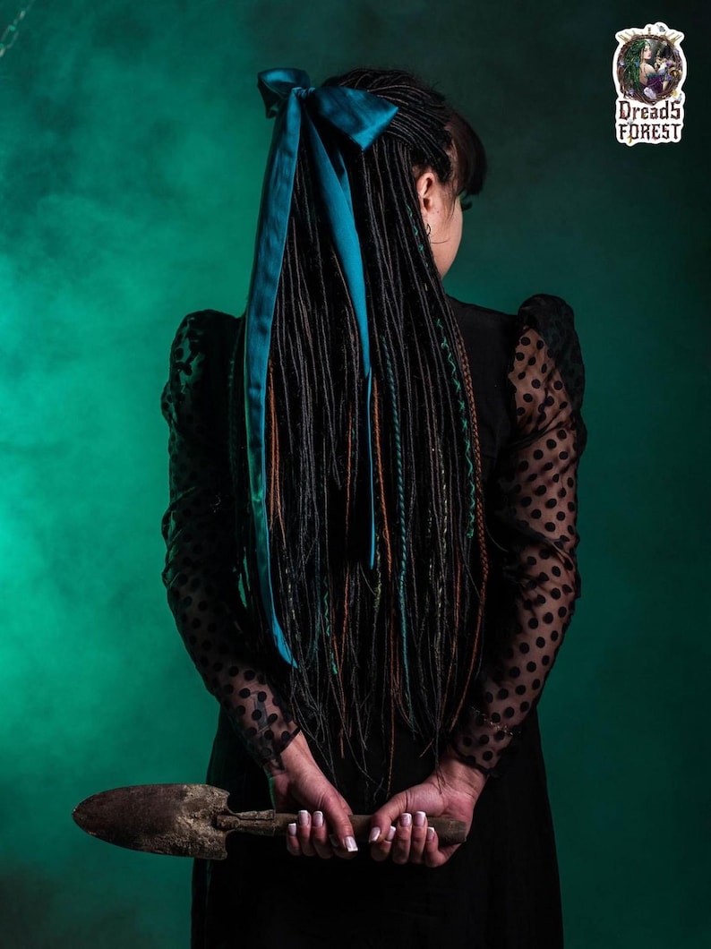 Handcrafted synthetic dreadlock extensions in forest green and black ombre. The set features a mix of thin twisted dreads, classic braids, and fishtail braids arranged in a fan shape. A model in a gothic setting holds a black skull, emphasizing a mystical forest witch aesthetic.