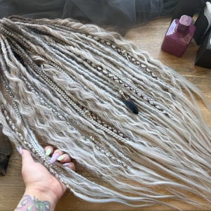 May include: A set of grey and silver dreadlocks, featuring a mix of wavy and straight sections. The dreads are decorated with small embellishments and a black charm. The image shows the dreadlocks on a wooden surface, with a logo in the top left corner.