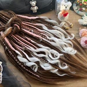 May include: Brown, white, and pink dreadlocks with decorative accents. The dreadlocks are styled with a mix of colors and textures, including wavy white strands and pink accents. The Dreads Forest logo is visible.