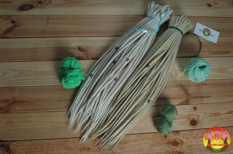 May include: Two bundles of white and beige wool dreadlocks with colorful beads. The dreadlocks are tied together with blue ribbon. Two balls of green yarn are on the wooden surface.