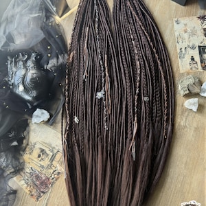 May include: A set of dark brown synthetic hair extensions featuring a mix of tight braids and loose, straight strands. The braids are adorned with small silver-toned metal charms and thin leather-like cord wraps, creating a textured appearance.