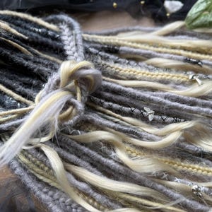 May include: Close-up of handmade dreadlocks in shades of gray, blonde, and cream. The dreads are adorned with small silver-colored charms and decorative stitching. The dreadlocks are tied together at the top.