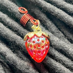 A close-up image of a red and gold strawberry-shaped charm with a spiral on top, resting on a textured gray background.