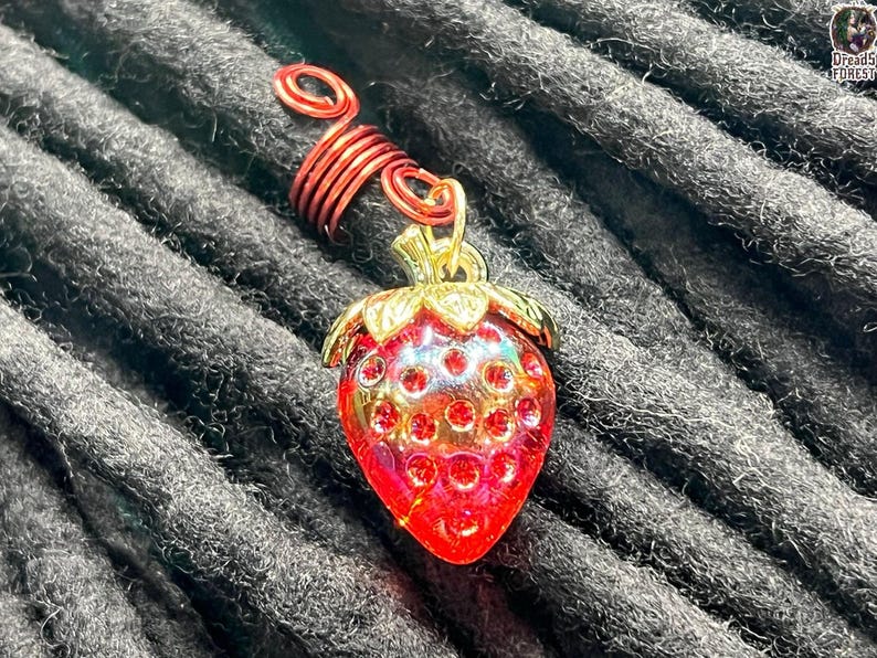 A close-up image of a red and gold strawberry-shaped charm with a spiral on top, resting on a textured gray background.