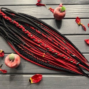 Puede incluir: Un conjunto de rastas sintéticas rojas y negras con detalles dorados y negros. Las rastas están dispuestas sobre una superficie de madera con manzanas rojas y pétalos de flores.