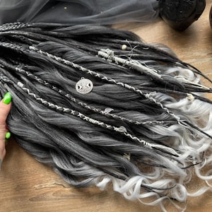 May include: A set of gray and white synthetic dreadlocks with decorative silver accents. The dreads are styled with intricate braids, charms, and white wavy ends. The image shows a close-up of the dreadlocks on a wooden surface.