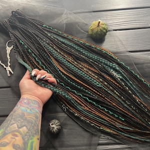 May include: A set of synthetic dreadlocks in black, brown, green, and gold colors. The dreadlocks are arranged in a fan shape and are lying on a black fabric surface.