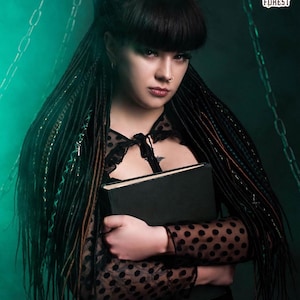 Close-up of a model holding a book, showing forest green and black synthetic dreads with cross-braided decorative wraps. The background features chains and green smoke, emphasizing a dark fantasy or urban witch look. The texture of the thin twisted dreads is clearly visible.