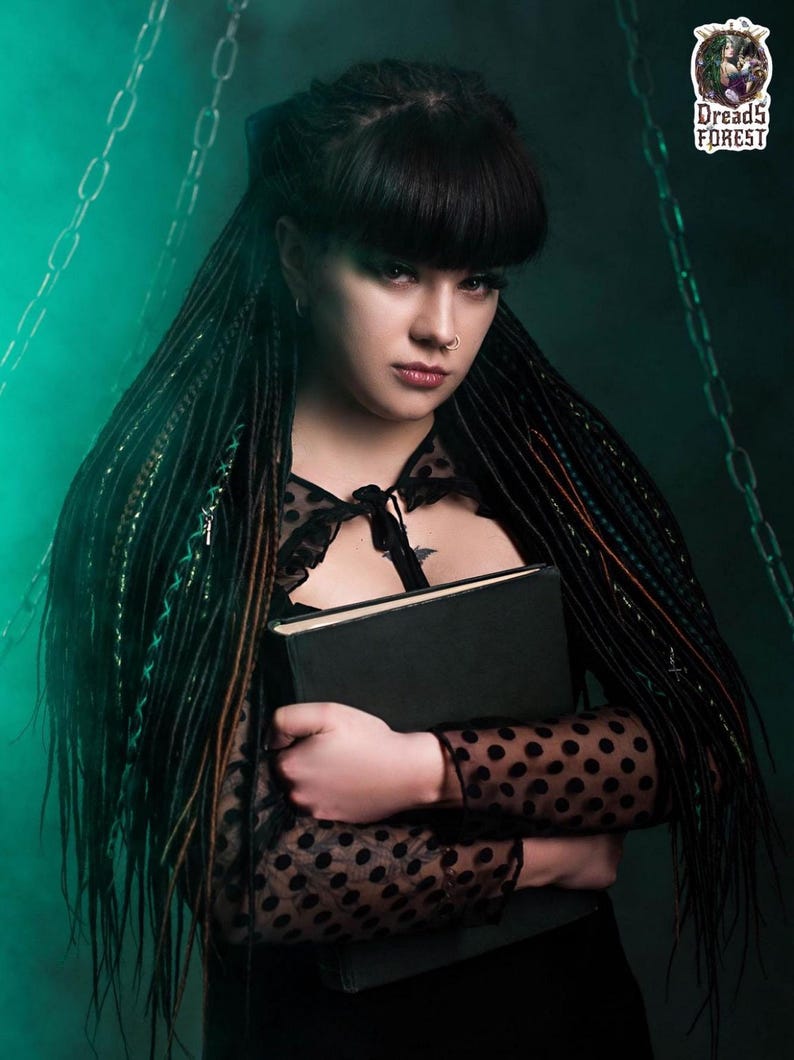 Close-up of a model holding a book, showing forest green and black synthetic dreads with cross-braided decorative wraps. The background features chains and green smoke, emphasizing a dark fantasy or urban witch look. The texture of the thin twisted dreads is clearly visible.