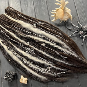 Puede incluir: Dreadlocks marrones oscuros y crema con detalles plateados. Los dreadlocks están colocados sobre una superficie de madera oscura, junto con una concha, una araña decorativa y un pequeño objeto en forma de panal.