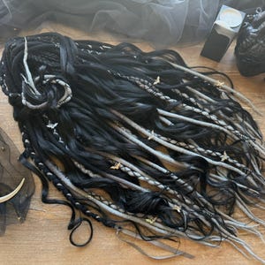 May include: Black and gray dreadlocks with silver accents and butterfly charms. The dreadlocks are styled with a decorative silver piece. The image also includes a logo, a crescent moon, and other decorative items.