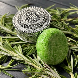 May include: A green rosemary shampoo bar with a silver decorative box on a dark wood surface with green rosemary sprigs.