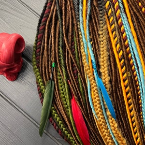 May include: A collection of synthetic dreadlocks in various shades of brown, yellow, red, and blue. The dreadlocks are decorated with feathers and beads.