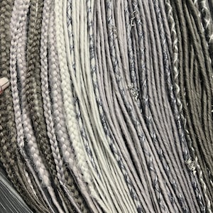 Puede incluir: Una colección de rastas sintéticas grises y blancas con abalorios plateados. Las rastas están dispuestas en fila y están listas para ser añadidas al cabello de una persona.