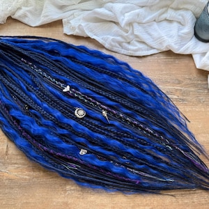 Könnte beinhalten: Ein Set aus blauen und schwarzen Dreadlocks mit dekorativen silbernen Charms und Perlen. Die Dreadlocks liegen auf einer Holzoberfläche, im Hintergrund ein weißes Tuch und eine Kerze in einem Glashalter. Das Dreads Forest Logo ist sichtbar.