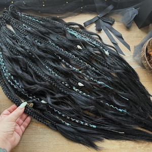 May include: Black dreadlocks with turquoise and white accents. The dreads are braided and decorated with small beads. A black perfume bottle and a bowl of nuts are also visible.