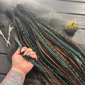 A hand holding a bundle of braided hair in various colors, including black, green, and brown, with a pair of scissors and a small green object on a wooden surface.