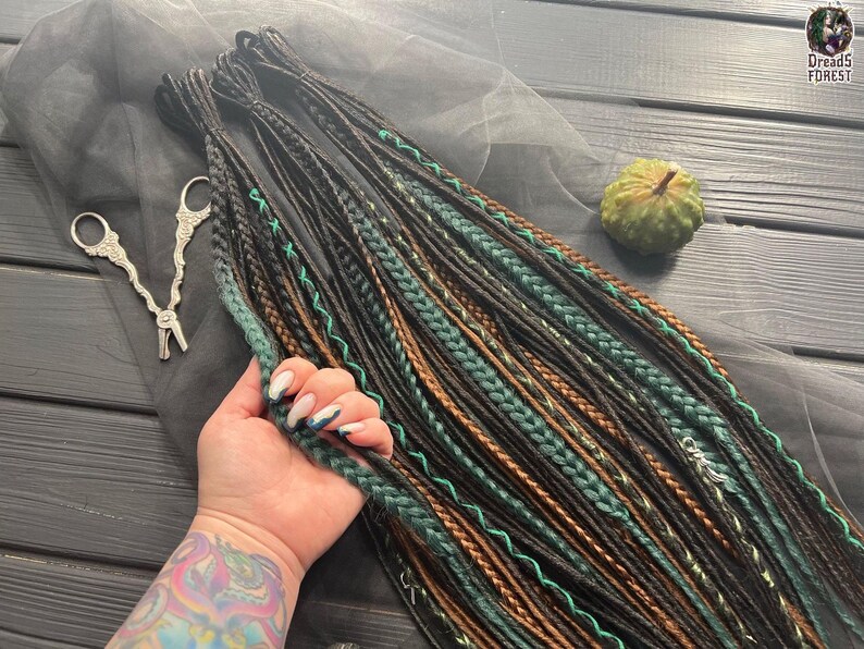 A hand holding a bundle of braided hair in various colors, including black, green, and brown, with a pair of scissors and a small green object on a wooden surface.