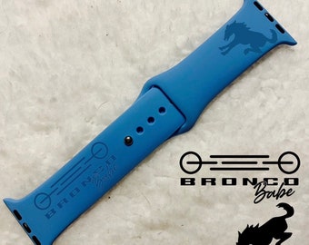 Peut inclure: Bracelet de montre Apple en silicone bleu avec un logo Bronco Babe noir. Le bracelet a une silhouette de cheval noir sur le dessus et le logo Bronco Babe sur le dessous.