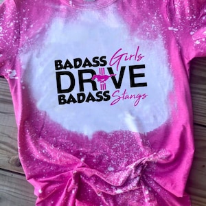 May include: Pink bleached t-shirt with a white bleach design. The design says "Badass Girls Drive Badass Stangs" with a pink Mustang logo.