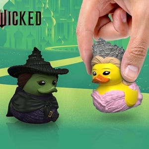 Op de afbeelding: Twee rubberen eendenbeeldjes geïnspireerd op de musical "Wicked". Eén eend is gekleed als de boze heks van het westen, in een zwarte hoed en cape. De andere eend wordt door een hand vastgehouden en is gekleed als Glinda, met een zilveren kroon en roze jurk.