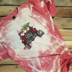 May include: Red and white bleached t-shirt featuring a graphic of a black Jeep with a red and black plaid design. The Jeep is decorated with a Christmas tree and gnomes, with the words "Merry Christmas" on the spare tire.