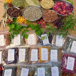 May include: A selection of dried herbs and flowers displayed in metal bowls and clear plastic bags. The herbs and flowers are in shades of green, brown, red, and yellow. Some bags have labels with text.
