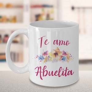 May include: White ceramic mug with the words "Te amo Abuelita" in pink script, accented by a watercolor floral design in shades of pink, yellow, and blue. The mug has a curved handle.