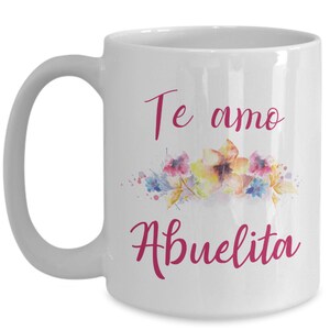 May include: White ceramic mug with a curved handle. The mug features the words "Te amo Abuelita" in a cursive, magenta font, along with a watercolor floral design in shades of pink, yellow, and blue.