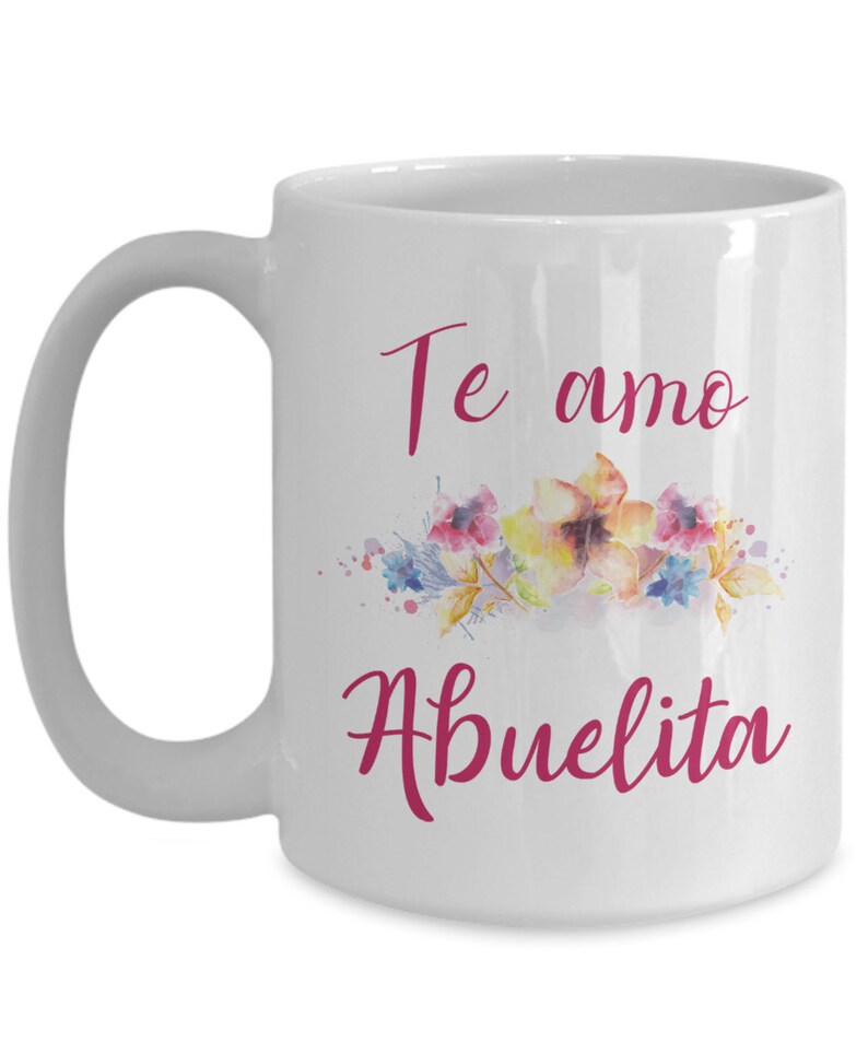 May include: White ceramic mug with a curved handle. The mug features the words "Te amo Abuelita" in a cursive, magenta font, along with a watercolor floral design in shades of pink, yellow, and blue.