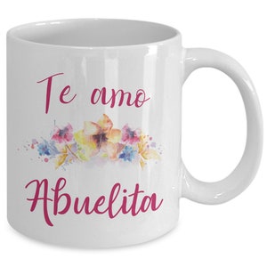 May include: White ceramic mug with a curved handle. The mug features the text "Te amo Abuelita" in pink script, above a watercolor floral design with yellow, pink, and blue flowers.