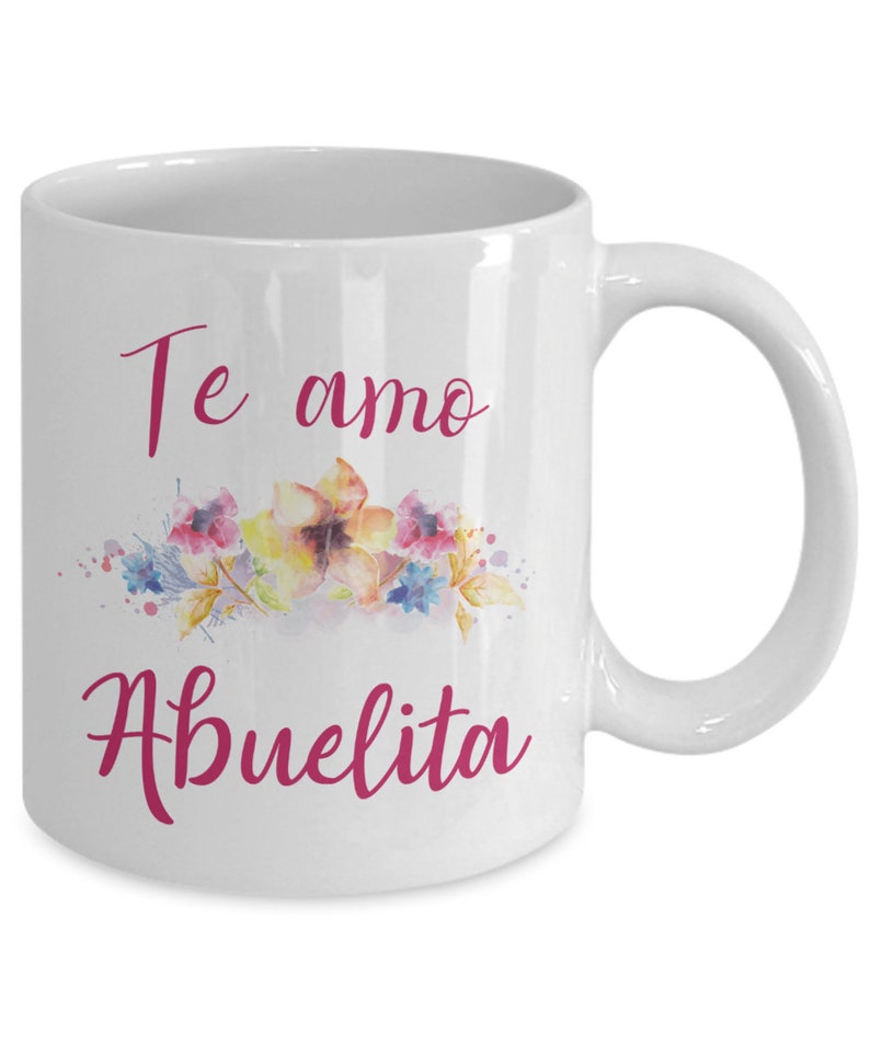 May include: White ceramic mug with a curved handle. The mug features the text "Te amo Abuelita" in pink script, above a watercolor floral design with yellow, pink, and blue flowers.