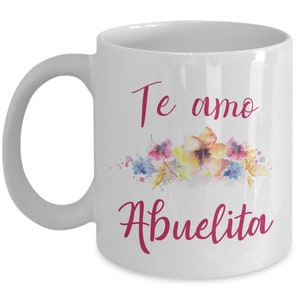 May include: White ceramic mug with a curved handle. The mug features the text "Te amo Abuelita" in pink script, along with a watercolor floral design in shades of pink, yellow, and blue.