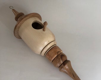 Puede incluir: Una casita de pájaros de madera con un acabado marrón claro y crema. La casita tiene forma de lágrima y tiene una pequeña abertura para que los pájaros puedan entrar. Tiene un pequeño pomo de madera en el lateral y un gancho de metal en la parte superior.