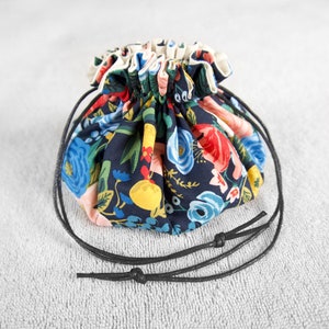 Op de afbeelding: Een klein trekkoordtasje met een zwarte bloemenprint en een zwarte trekkoordsluiting. De tas is gemaakt van stof en heeft een geplooide bovenkant.
