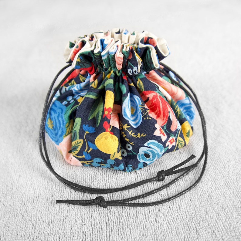 Op de afbeelding: Een klein trekkoordtasje met een zwarte bloemenprint en een zwarte trekkoordsluiting. De tas is gemaakt van stof en heeft een geplooide bovenkant.