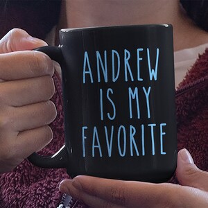May include: Black coffee mug with a white handle. The mug has blue text that reads "ANDREW IS MY FAVORITE".