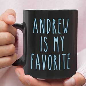 May include: Black coffee mug with white handle. The mug has blue text that reads "Andrew is my favorite".