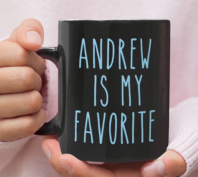 May include: Black coffee mug with white handle. The mug has blue text that reads "Andrew is my favorite".