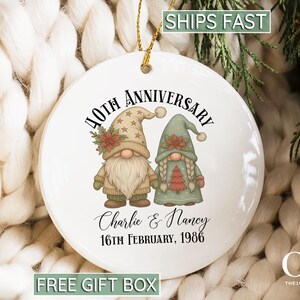 May include: A round white ceramic ornament featuring a graphic of two gnomes with poinsettias on their hats. The text reads 40TH ANNIVERSARY, Charlie & Nancy, and 16TH FEBRUARY, 1986. The ornament hangs from a gold string against a white knit background.