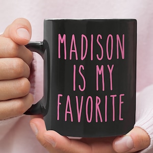 May include: Black coffee mug with a pink inscription that reads "MADISON IS MY FAVORITE".