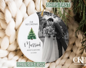 May include: A round ceramic Christmas ornament featuring a split design. One side displays a black and white photograph of a couple, while the other side is white with a green Christmas tree illustration and the text: OUR FIRST CHRISTMAS Married ASHLEY & MICHAEL 4.5.2026. It hangs from a gold string.