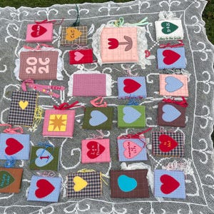 Op de afbeelding: Een verzameling kleine, vierkante stoffen items met diverse ontwerpen, waaronder harten, sterren en bloempatronen. Sommige items bevatten tekst zoals "LOVE" en namen. De items zijn gerangschikt op een wit kanten tafelkleed.